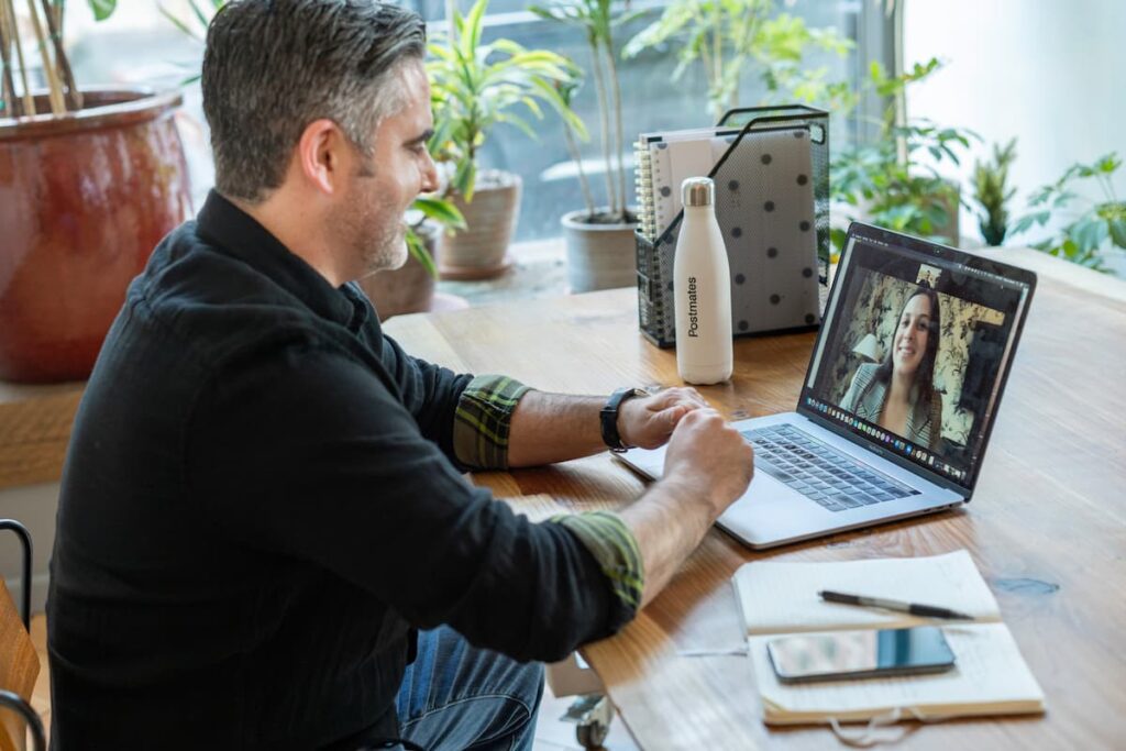 A person sitting at a desk with a laptop open. On the laptop screen is another person. They are having a video conference call.