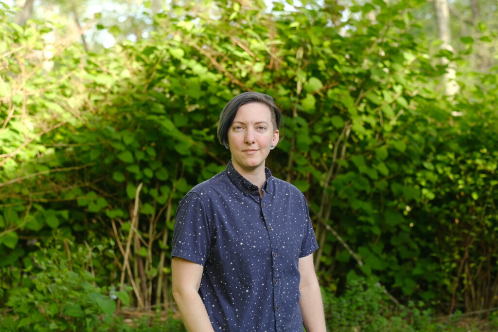 A photograph of Logan from the waist up wearing a dark blue short sleeved button up shirt standing in front of a swath of greenery.